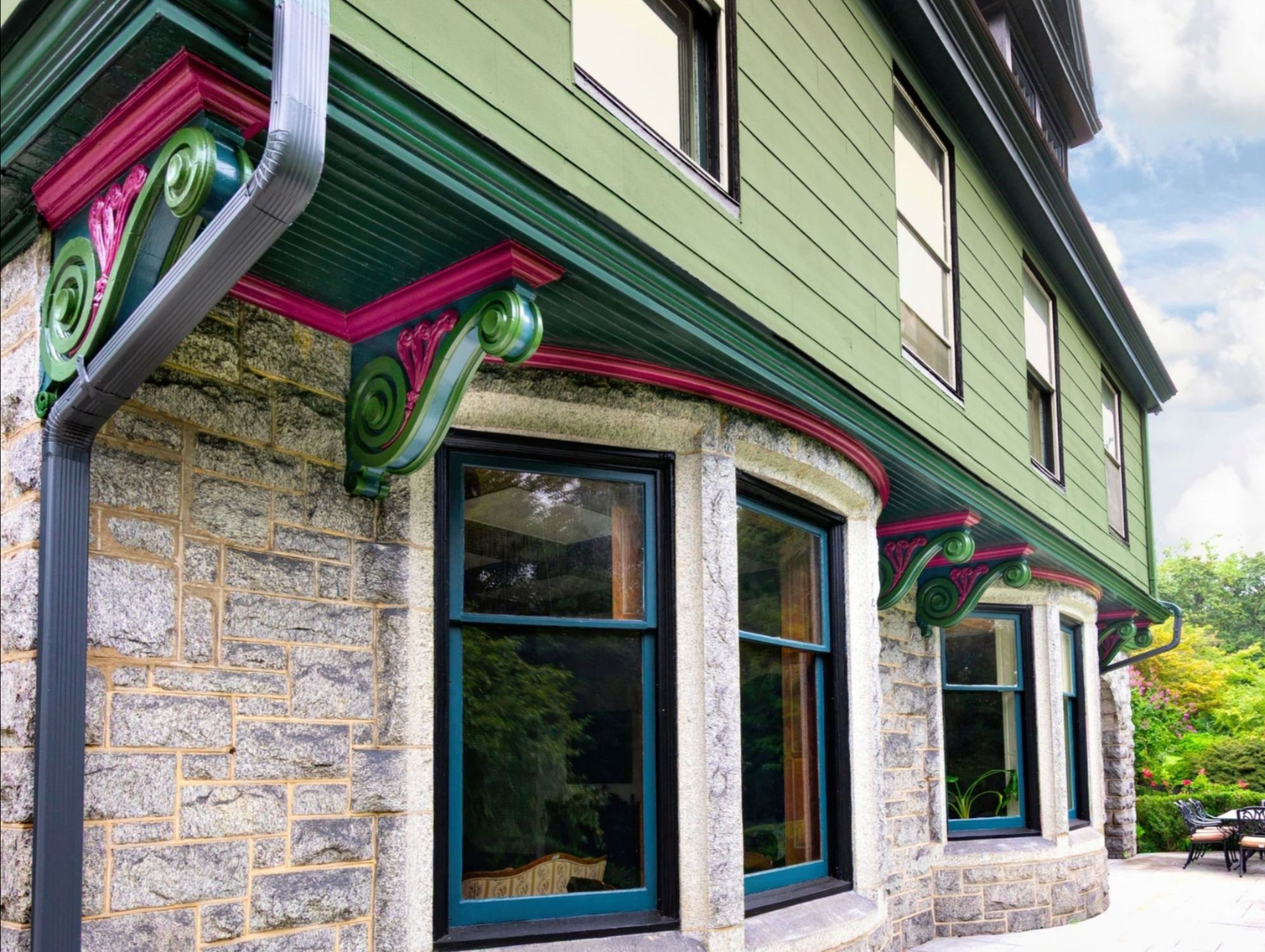 Nolan Painting Company- Havertown, PA Corner of a stone building with ornate green and magenta brackets under a green roof, blue-trimmed windows, and a garden area visible nearby.