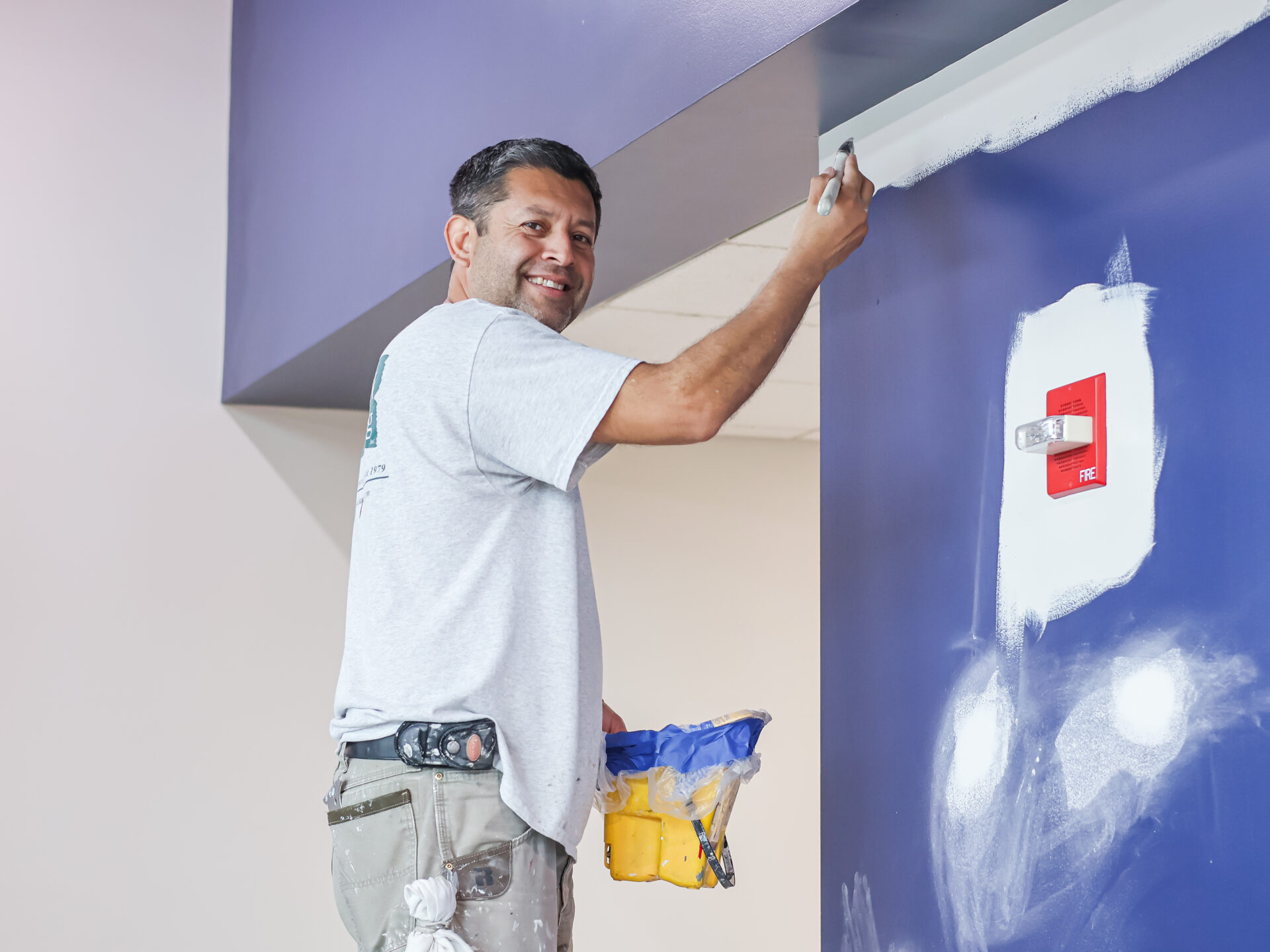 Smiling painter in a gray shirt and tool belt applies white paint to a blue wall.