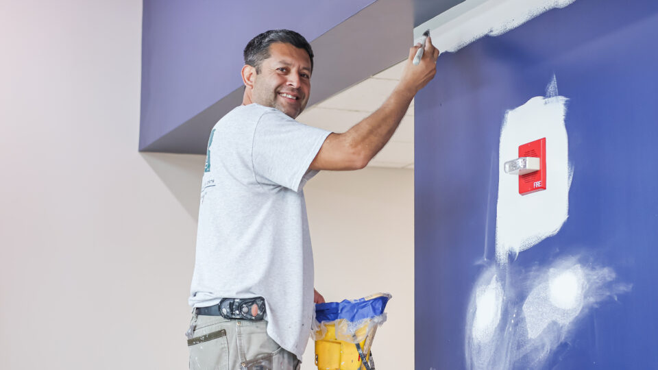 Smiling painter in a gray shirt and tool belt applies white paint to a blue wall.
