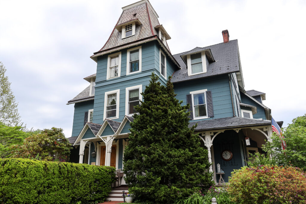 Blue Victorian house with a tall turret, slate roof, white-trimmed windows, and lush front garden.
