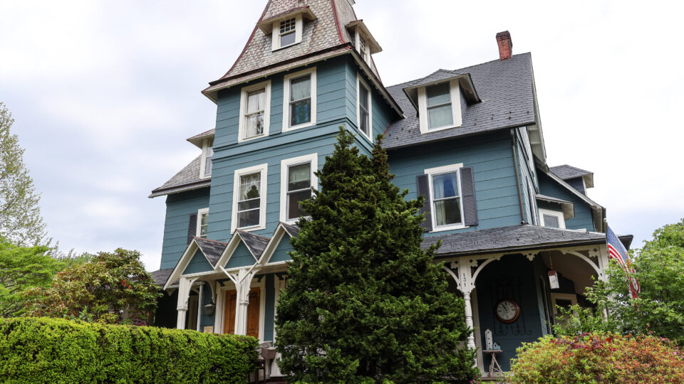 Blue Victorian house with a tall turret, slate roof, white-trimmed windows, and lush front garden.