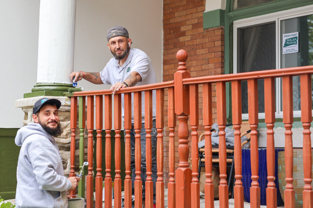 Two painters on a porch rail, one using a roller to paint a red railing while the other looks on wearing a gray hoodie and cap.