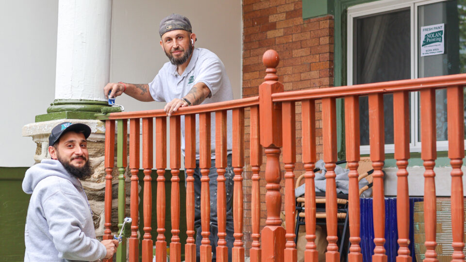 Two painters on a porch rail, one using a roller to paint a red railing while the other looks on wearing a gray hoodie and cap.