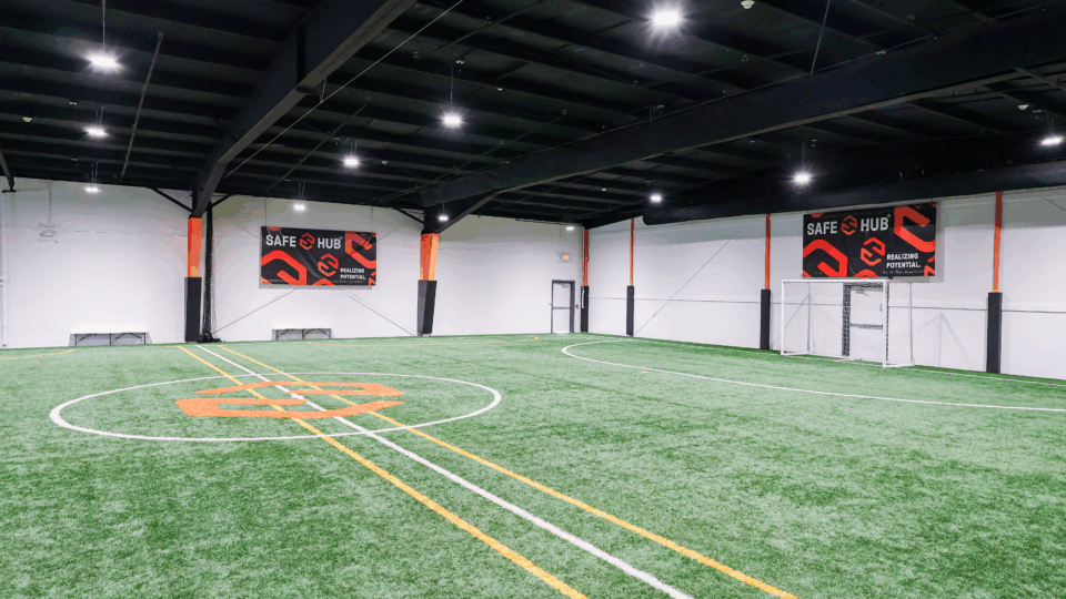 Indoor sports facility with artificial turf field, white and yellow lines, and banners reading Safe Hub along white walls.