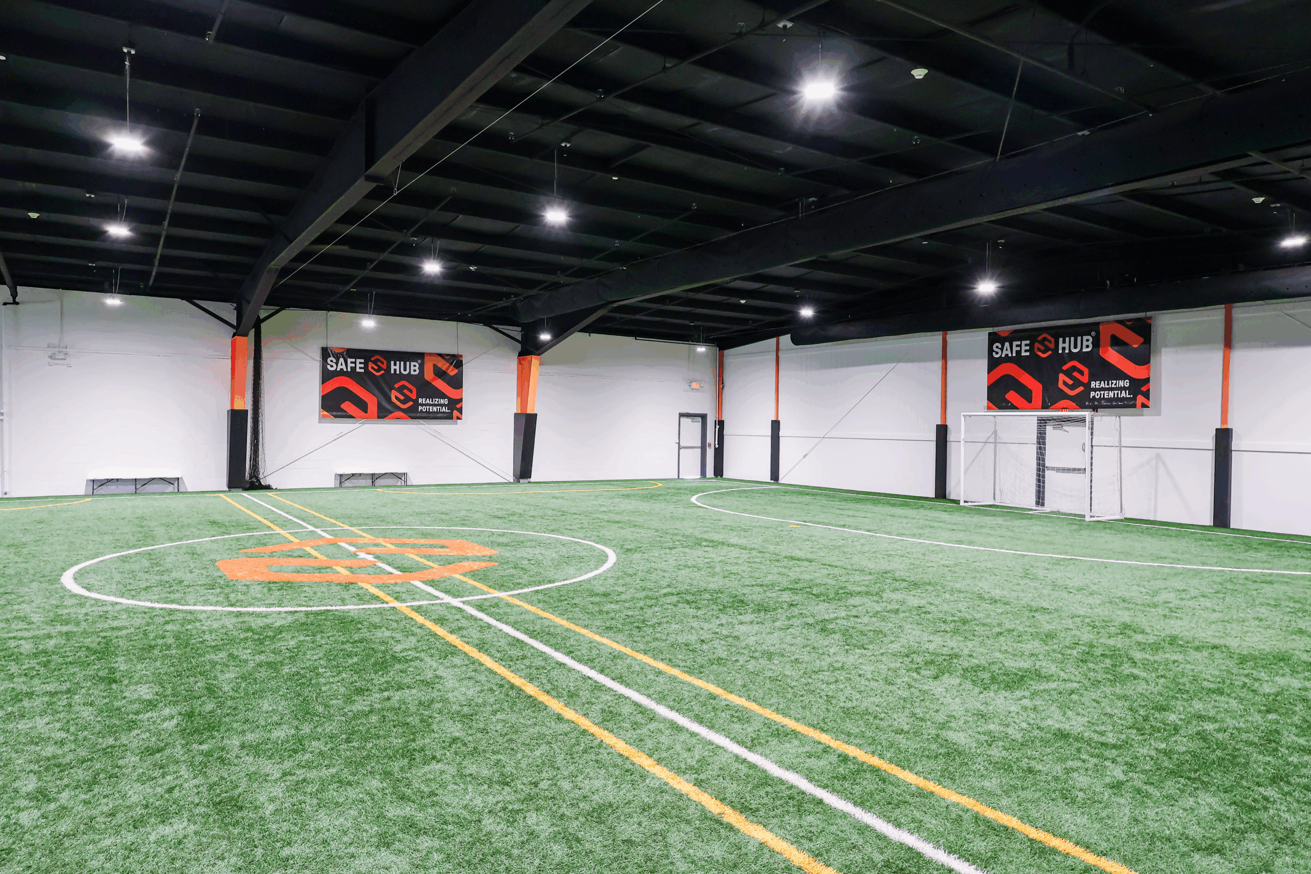 Indoor sports facility with artificial turf field, white and yellow lines, and banners reading Safe Hub along white walls.