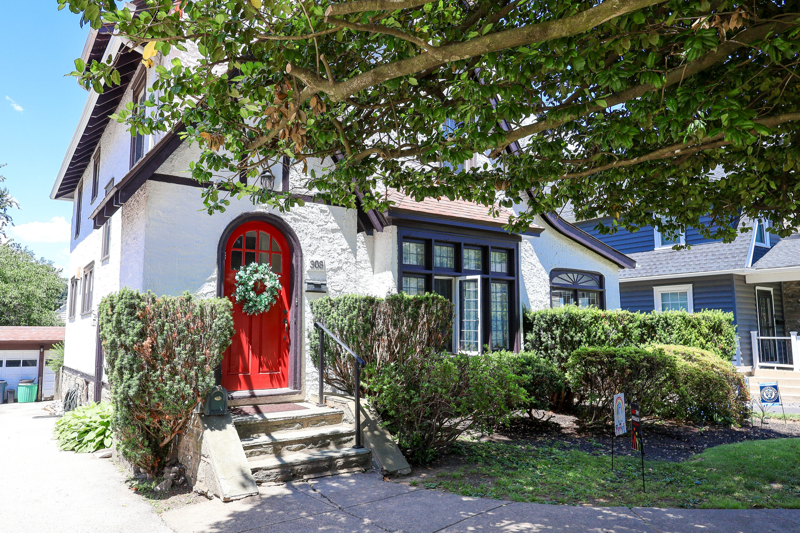 White stucco house with a bright red arched front door beneath a large leafy tree, with shrubs along the walkway.