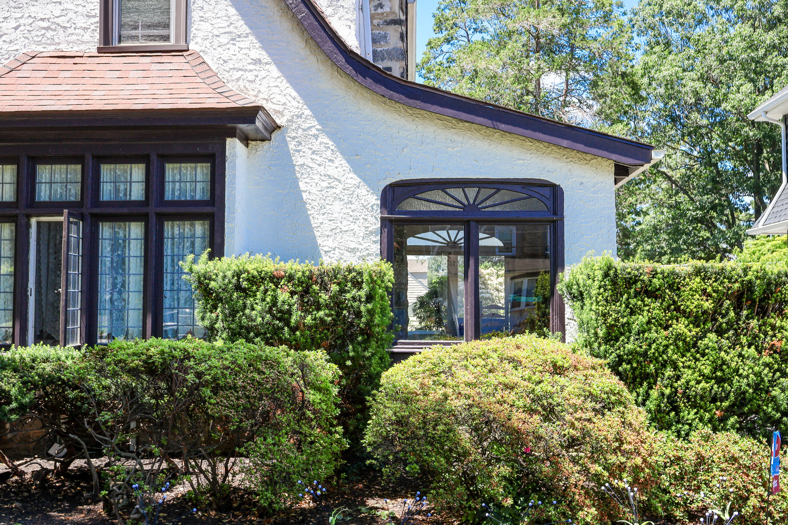 White stucco house with dark purple trim and arched window, surrounded by green hedges and trees in bright daylight