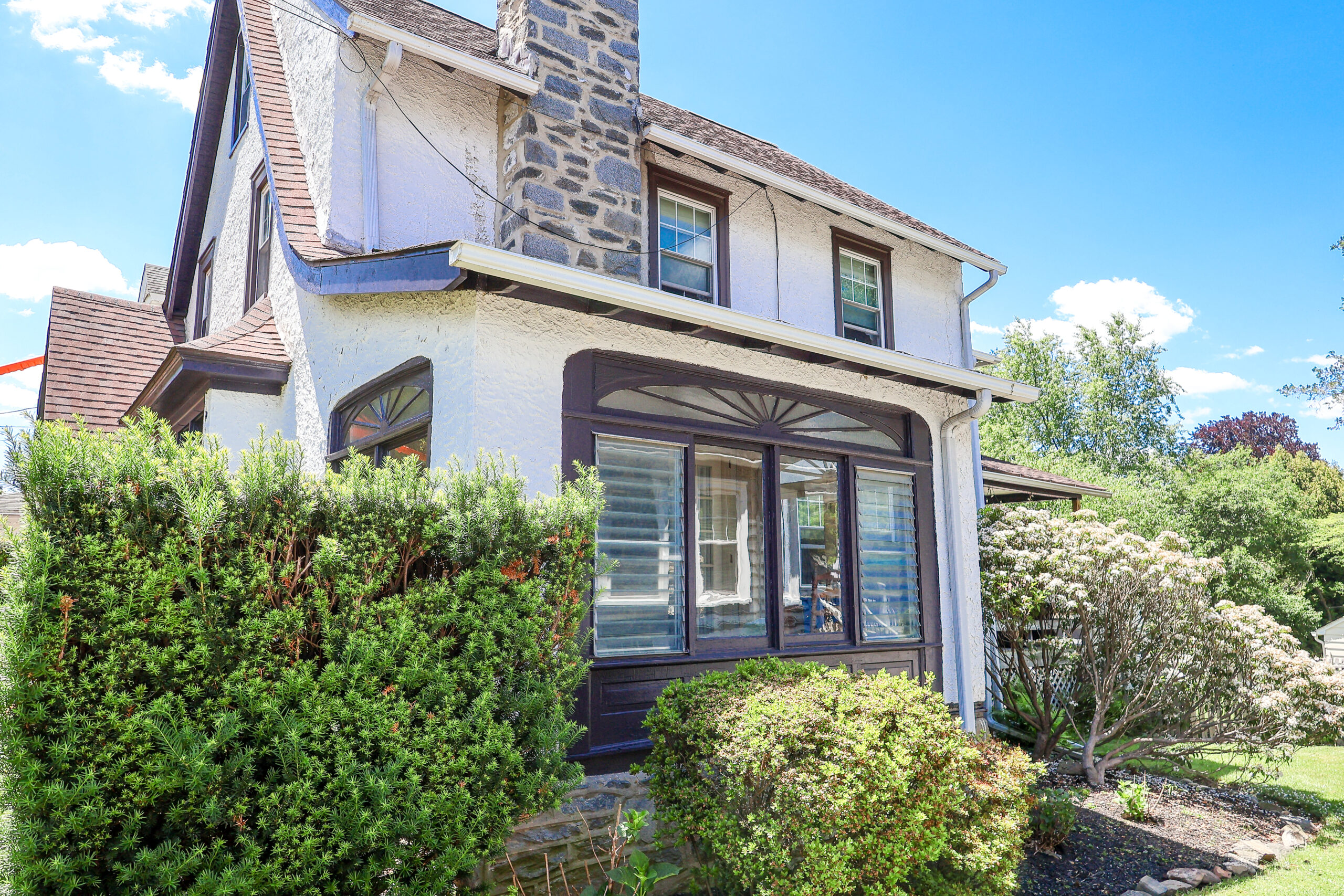 Two-story house with white stucco walls, a stone chimney, brown trim, and large front windows, surrounded by dense green shrubs under a blue sky.