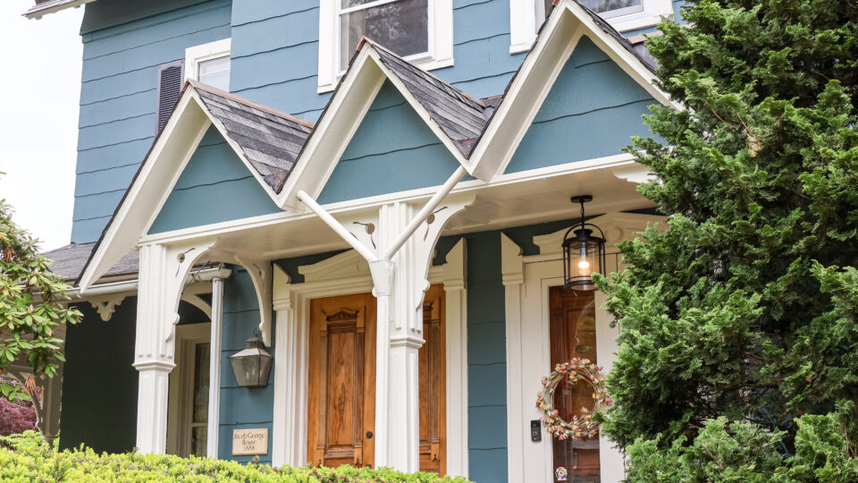 Blue two-story house with decorative white trim and three steep gabled dormers over the porch, surrounded by green shrubs and a wooden front door.
