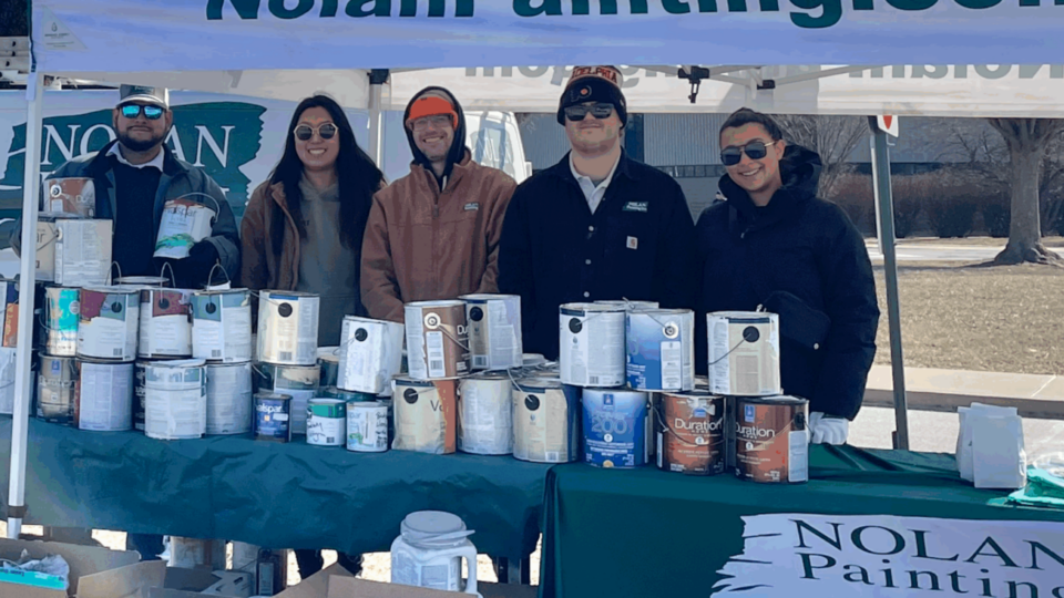 Group of five people standing under a Nolan Painting tent at an outdoor event, with paint cans on the table and banners in the background.