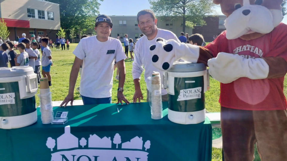 Mascot of a large cartoon dog posing with two men at an outdoor community event, a table with Nolan Painting branding in the foreground.