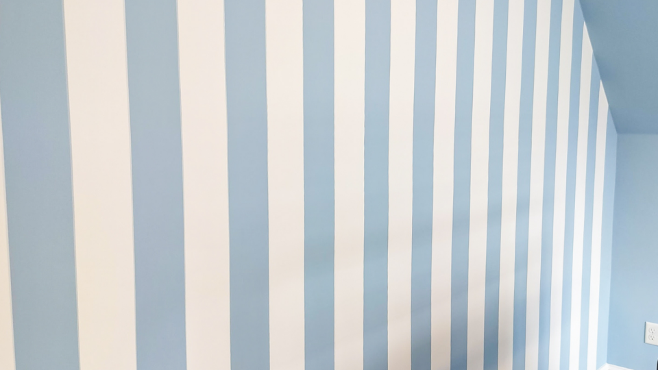 Interior wall with alternating light blue and white vertical stripes in a corner of a room with carpeted floor.