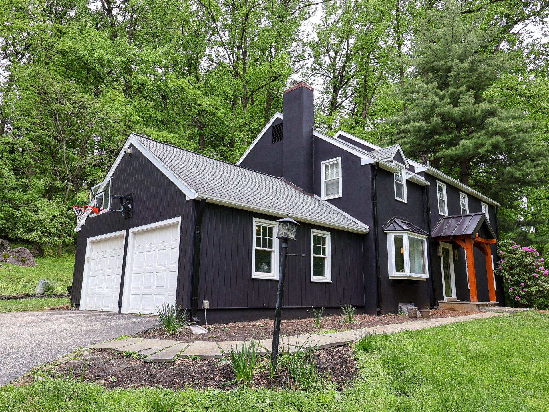 Dark-gray/blue house with white trim and a two-car garage, basketball hoop mounted on the left, and a lush green yard and trees in the background.