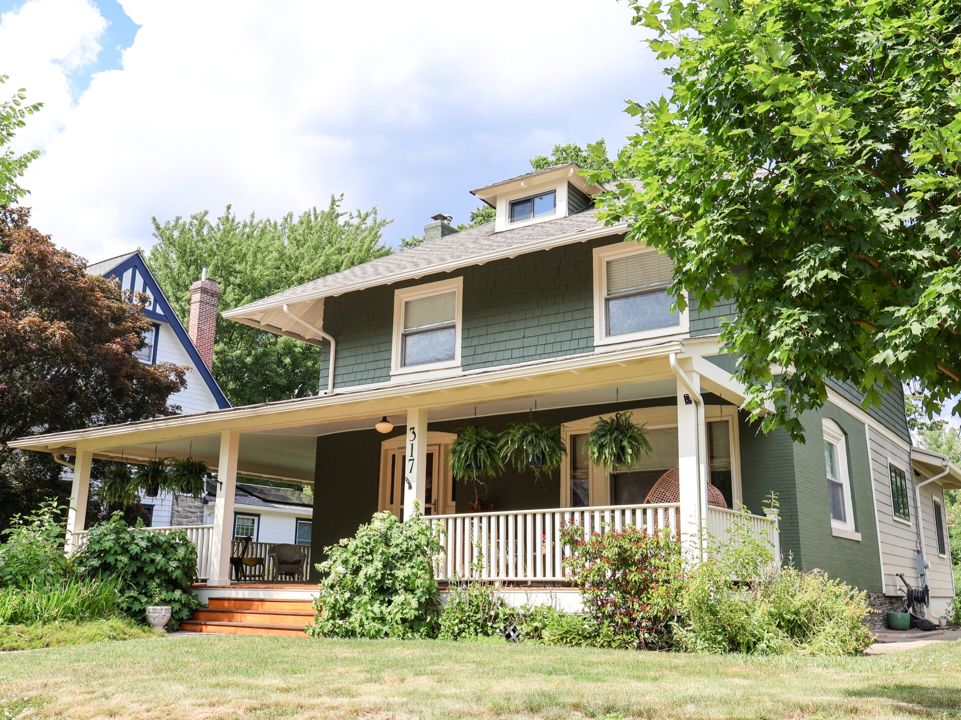 Two-story green house with white trim and a front porch, surrounded by trees and a bright blue sky.