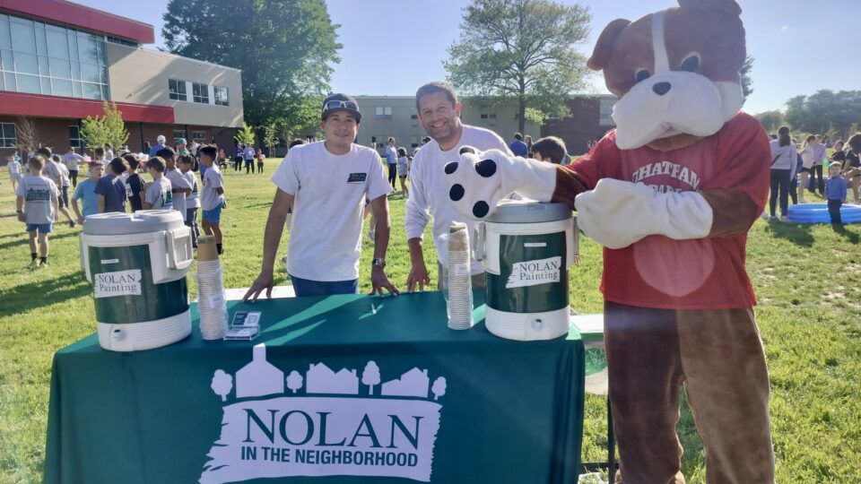 Mascot dog in a red shirt greets volunteers at a Nolan Painting community event with paint cans on a table and a banner nearby.