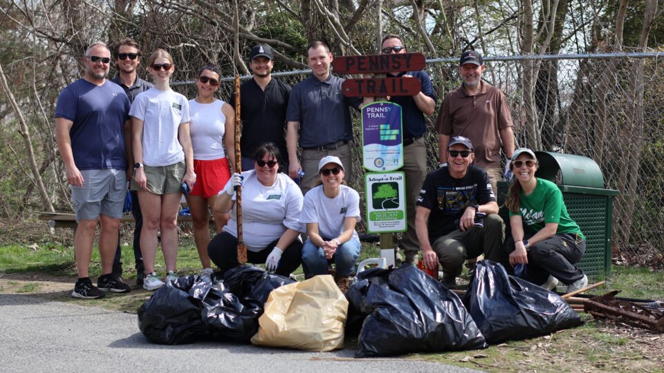 Group of volunteers posing outdoors behind a pile of large garbage bags and a Pennsy Trail sign.
