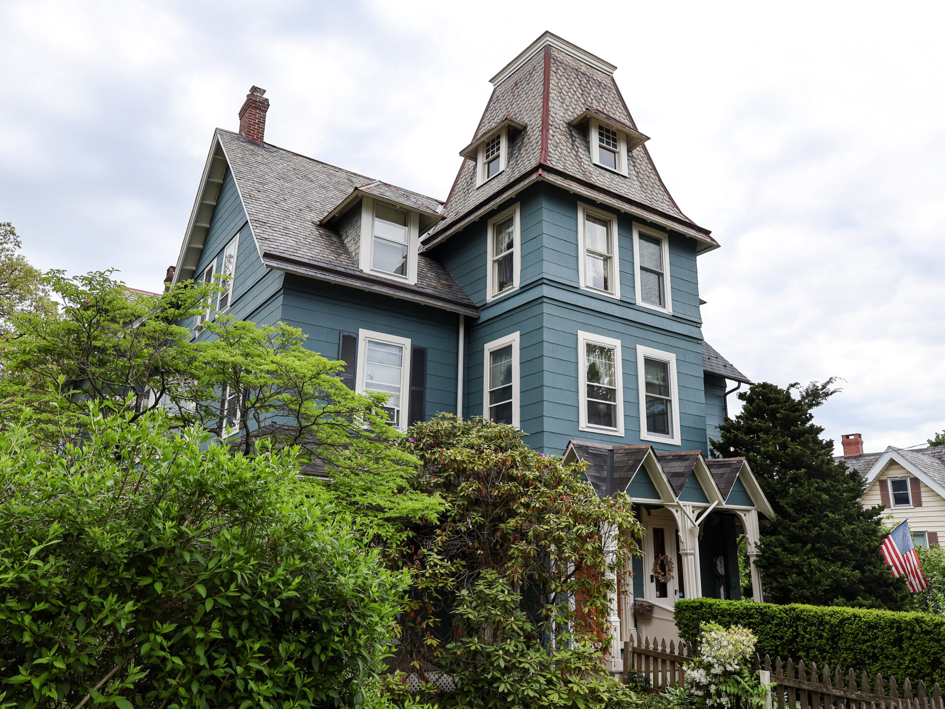 Blue Victorian house with steep slate roof and dormer windows, surrounded by green shrubs and a wooden fence under a cloudy sky