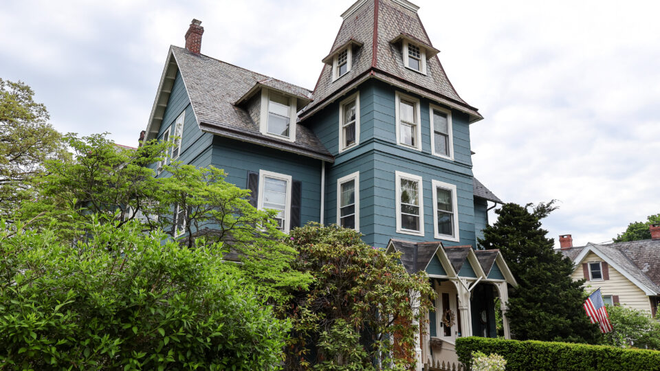 Blue Victorian house with steep slate roof and dormer windows, surrounded by green shrubs and a wooden fence under a cloudy sky