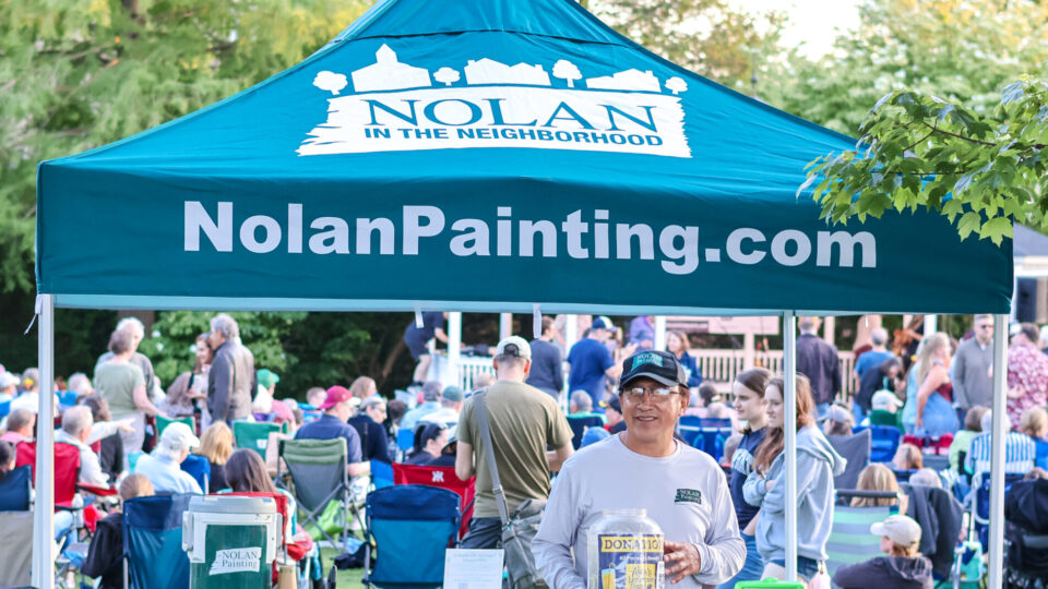 Outdoor community event under a blue Nolan Painting tent, with a crowd of people seated on lawn chairs in a park and volunteers at the booth nearby.