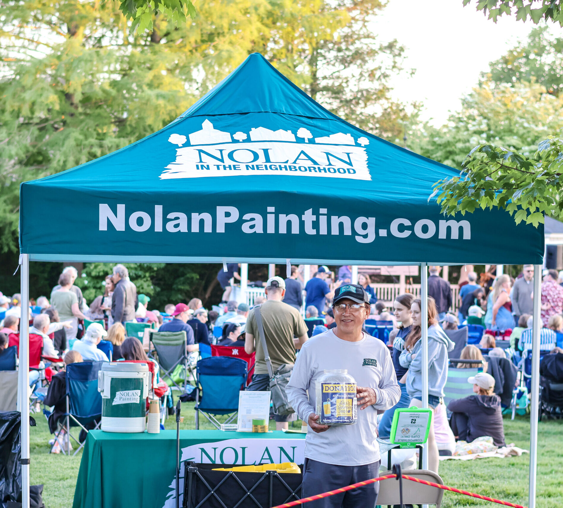 Outdoor community event under a blue Nolan Painting tent, with a crowd of people seated on lawn chairs in a park and volunteers at the booth nearby.