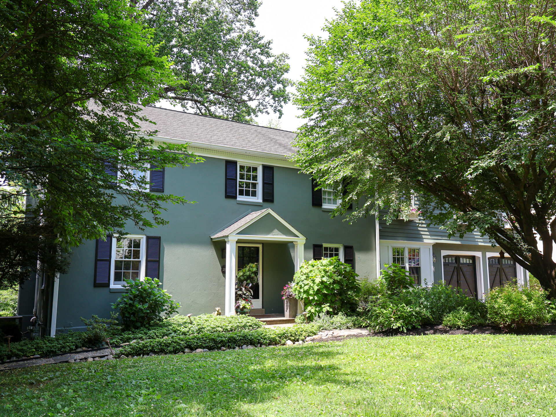 Green two-story house partially shaded by large trees, with front porch and garden area.