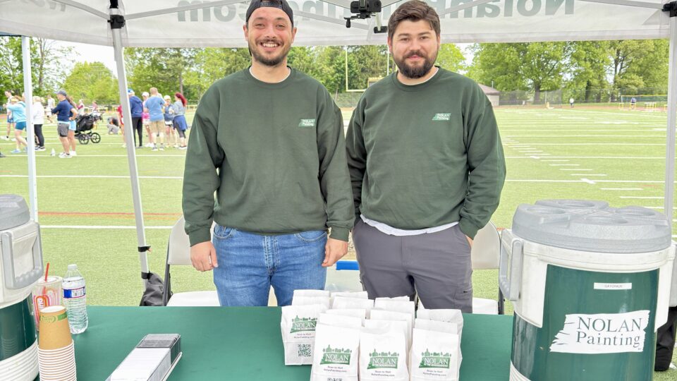 Two men in green shirts stand behind a Nolan Painting booth under a white canopy at an outdoor community event on a sports field, smiling for the camera.