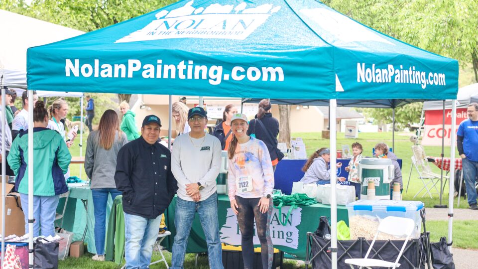 Group of volunteers under a blue Nolan Painting tent at an outdoor event.