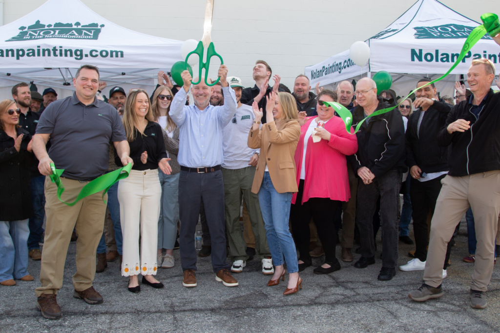 Group of people at a ribbon-cutting event, a man holds oversized green scissors while a green ribbon unfurls, Nolan Painting tents behind.