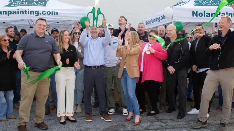 Group of people at a ribbon-cutting event, a man holds oversized green scissors while a green ribbon unfurls, Nolan Painting tents behind.
