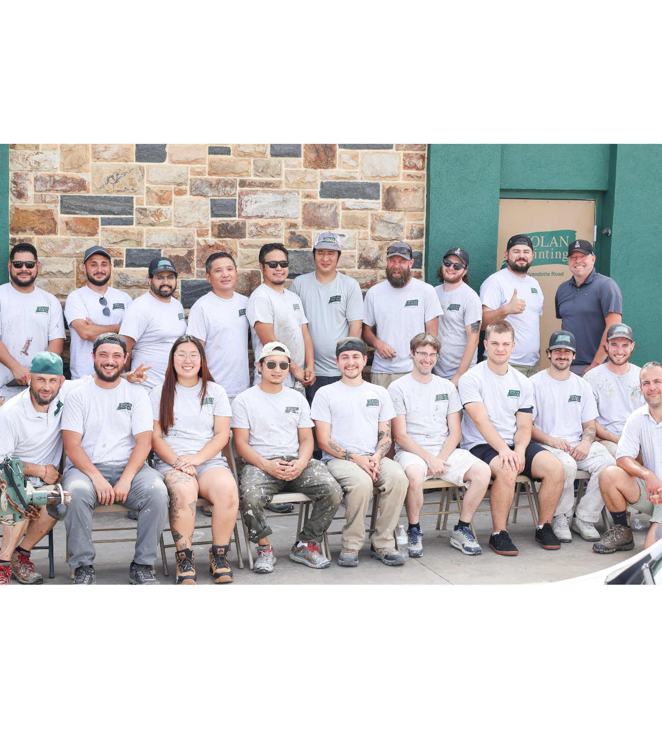 Group of workers posing for a team photo in matching gray shirts outside a stone wall building.