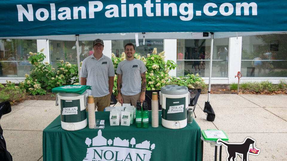 Two men in gray Nolan Painting shirts stand behind a promotional booth under a blue NolanPainting canopy.
