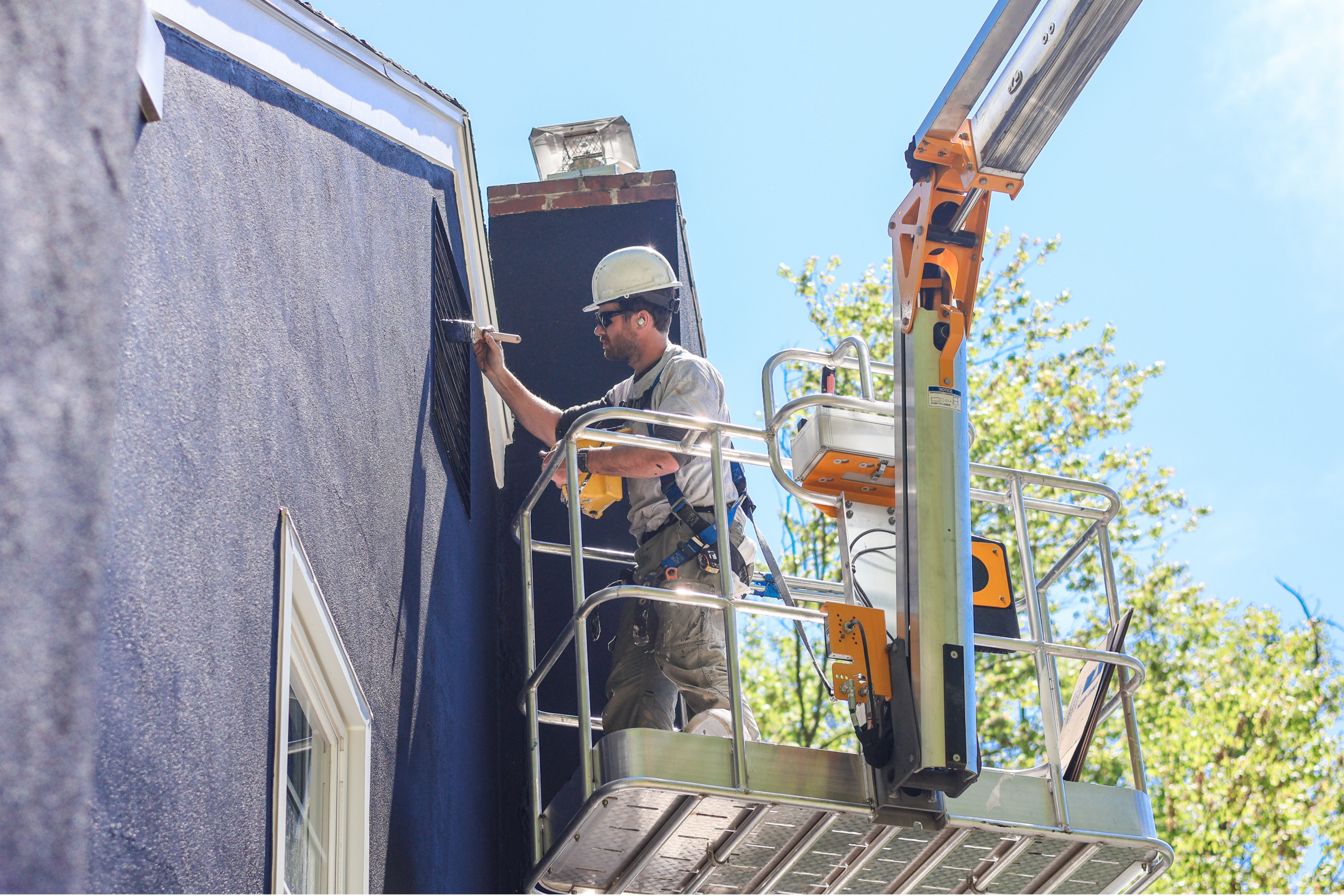 Nolan Painting Company- Havertown, PA Construction worker in a hard hat on a raised platform painting a blue exterior wall with a paint roller
