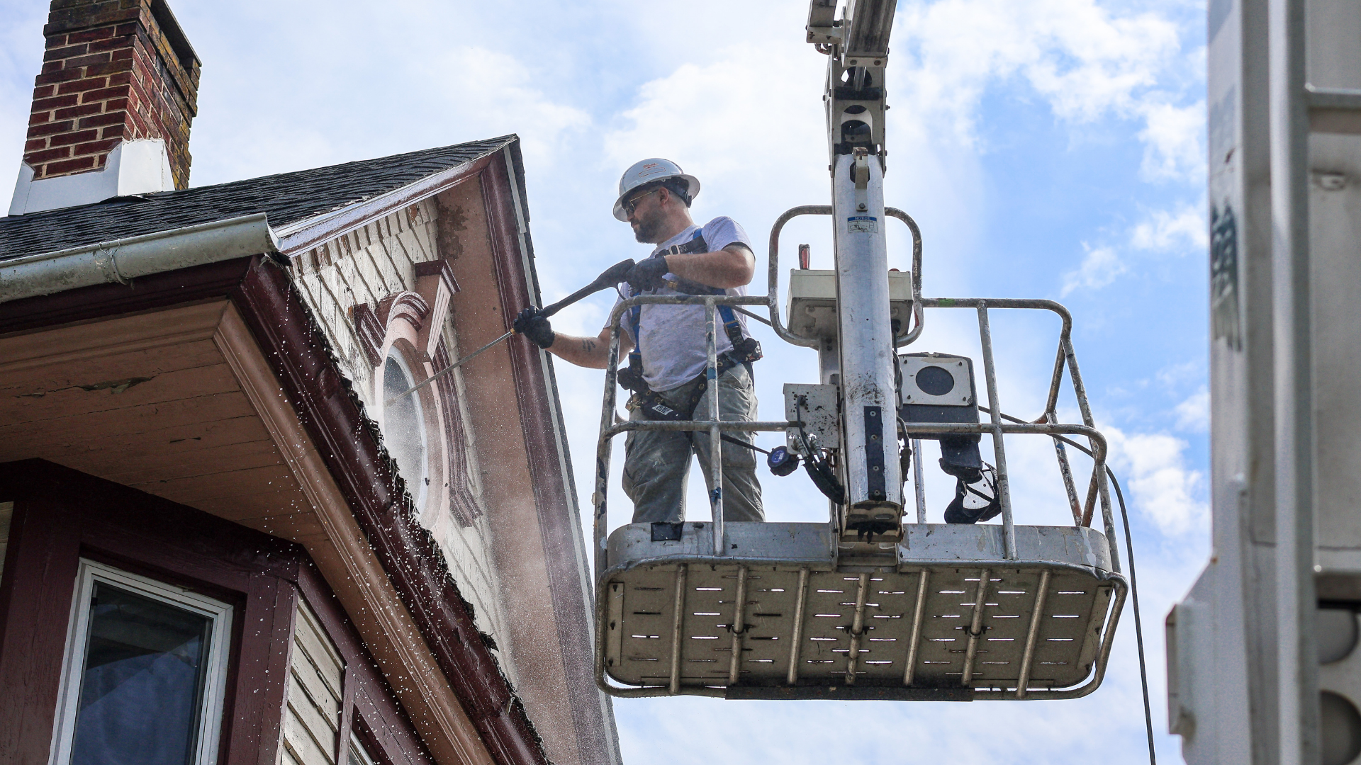 Nolan Painting Company- Havertown, PA Worker in a helmet on a raised lift power-washing the window of a brick house against a blue sky.