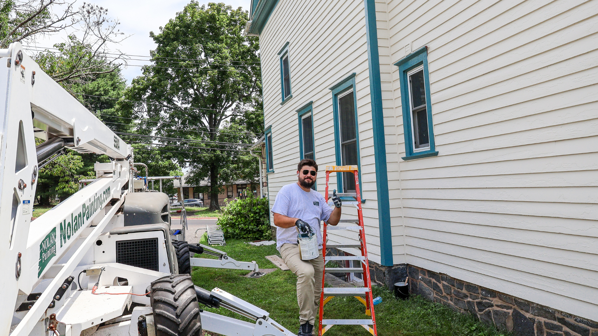 Nolan Painting Company- Havertown, PA A painter wearing gloves and sunglasses stands on a red ladder beside a beige house with blue trim, holding a paint can and brush while a Nolan Painting truck is nearby.