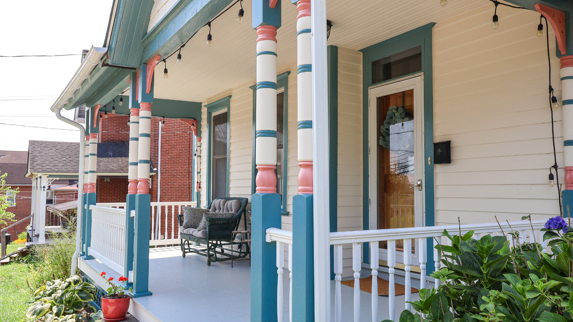 Nolan Painting Company- Havertown, PA Front porch of a beige house with teal trim and pastel striped columns, wicker seating, and string lights. The railing and plants line the porch edge.