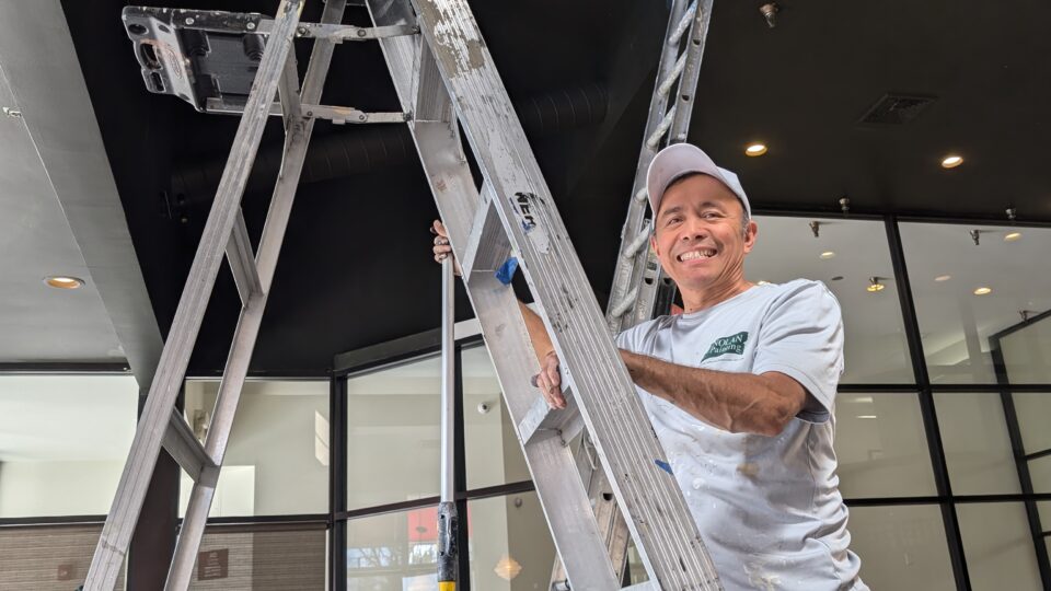 A smiling painter stands on a tall ladder, painting a dark ceiling with exposed ductwork above him inside a showroom or office space.