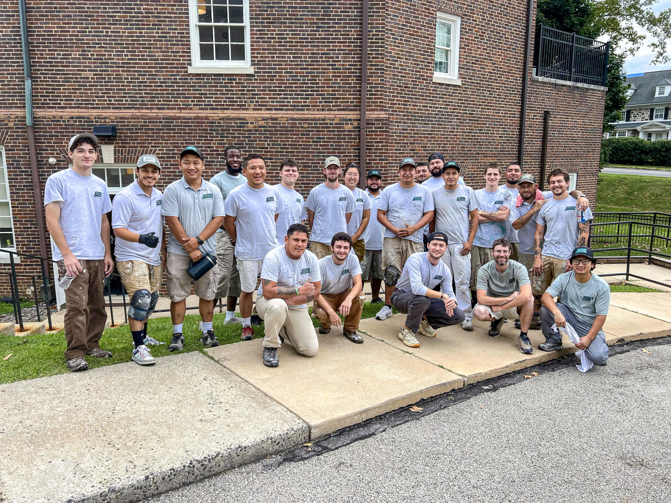 Nolan Painting Company- Havertown, PA Group of volunteers in gray shirts posing together outside a brick building, some kneeling and smiling.