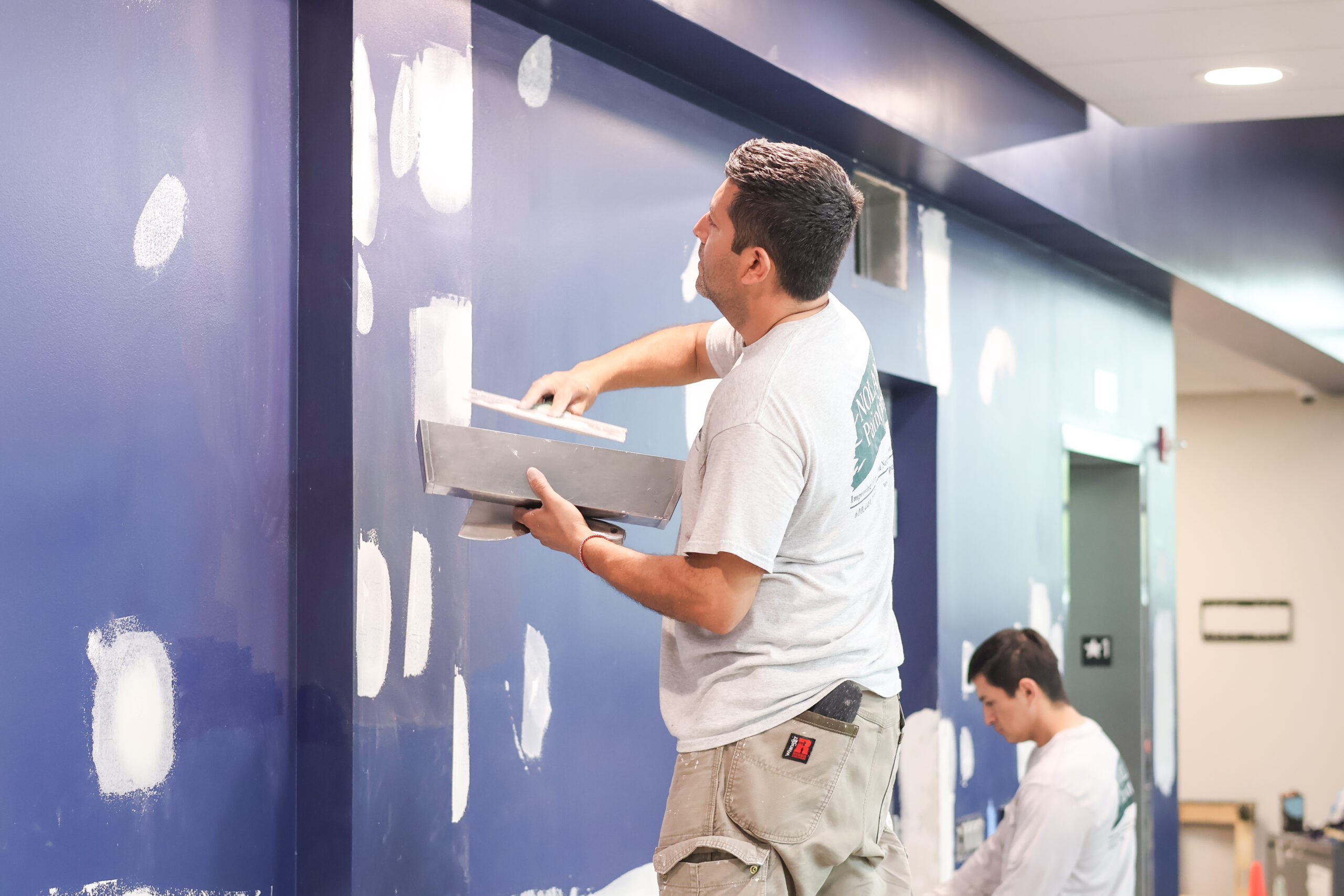 Nolan Painting Company- Havertown, PA Man in a gray shirt and tan shorts roller-paints a blue wall, creating white patches in a hallway or classroom setting
