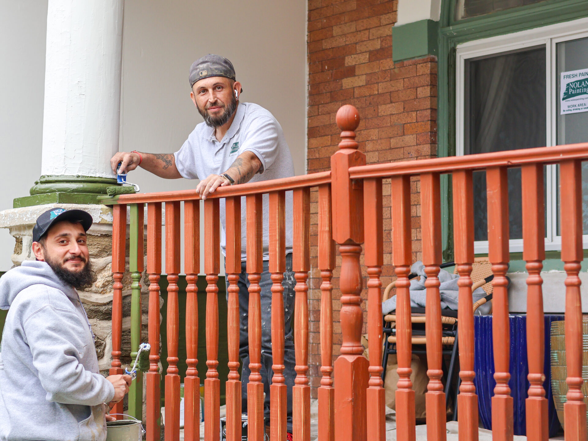 Two men painting a wooden porch railing orange, one on the railing with a paint roller and the other standing nearby wearing a gray hoodie and cap.