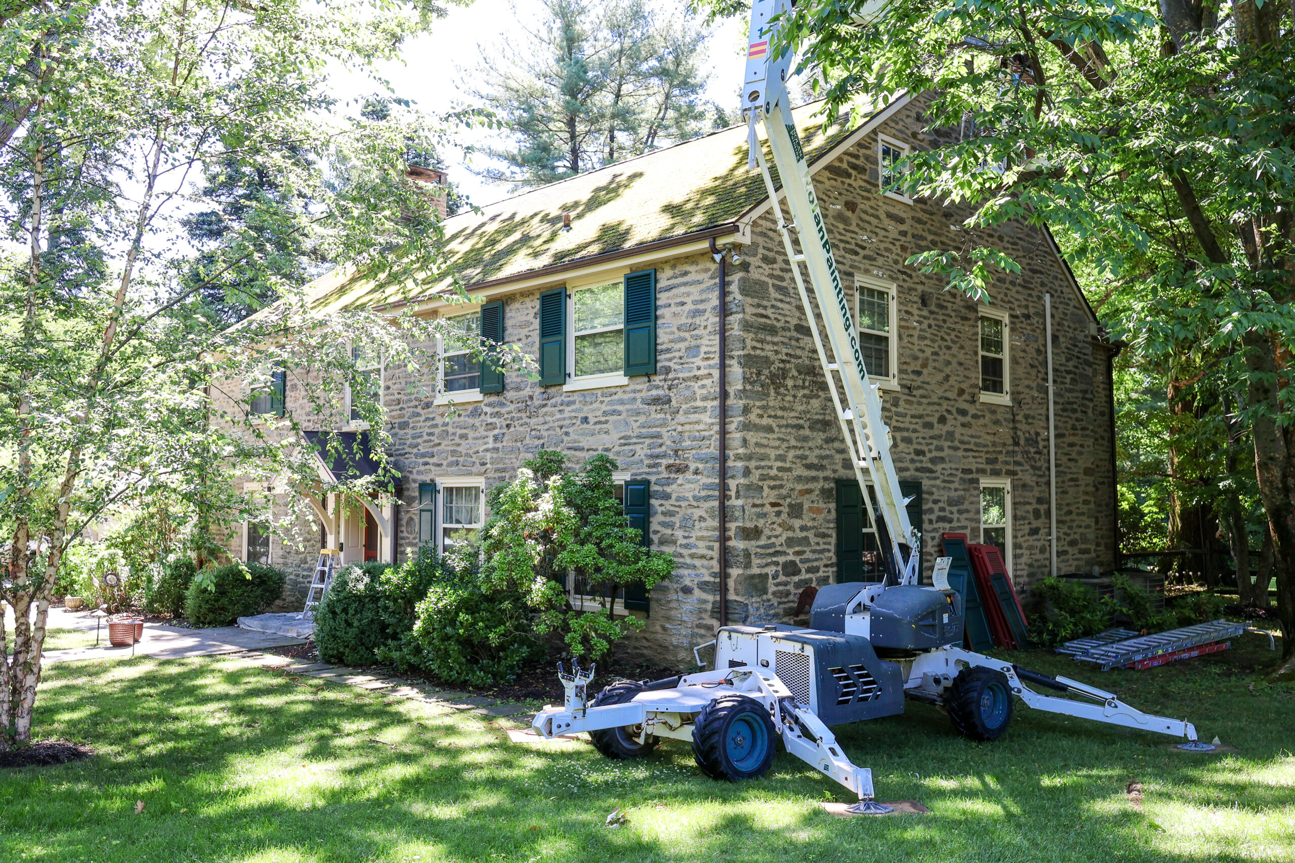 Stone cottage with green shutters, surrounded by trees; a boom lift and ladders in the yard for exterior work.