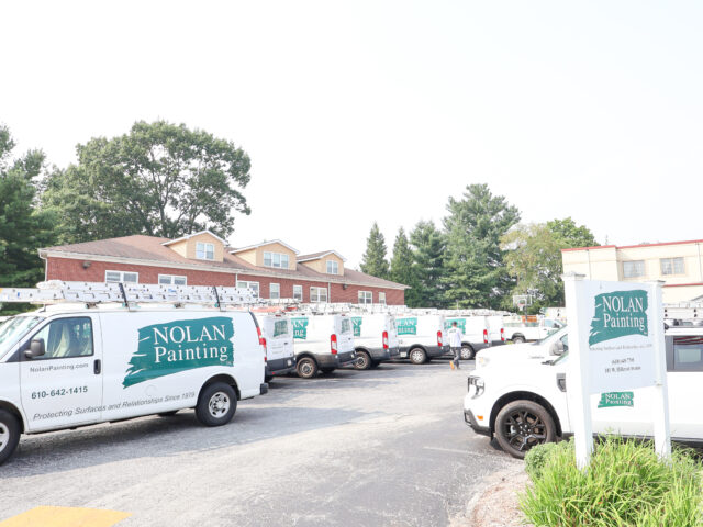 Fleet of Nolan Painting vans with ladders on top parked in a lot, company signage visible on the sides of the vehicles.