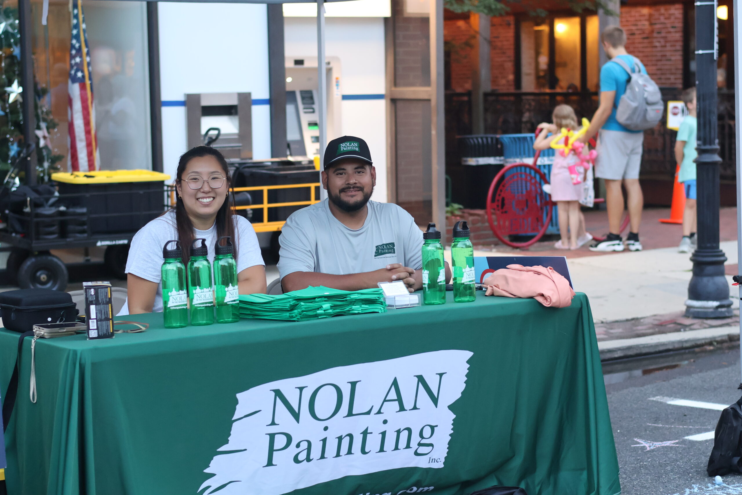 Two smiling people sit at a booth with a green tablecloth labeled Nolan Painting Inc., water bottles and flyers visible. Behind them is a city street scene.