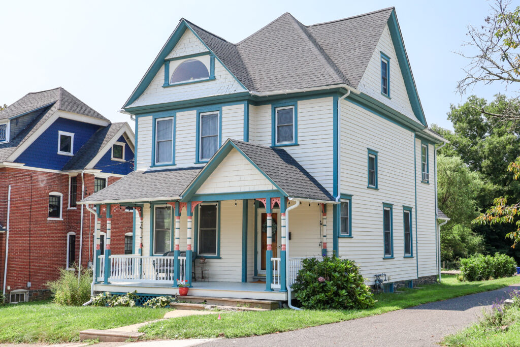 Two-story cream house with teal trim and wraparound porch beside a brick home on a sunny street.