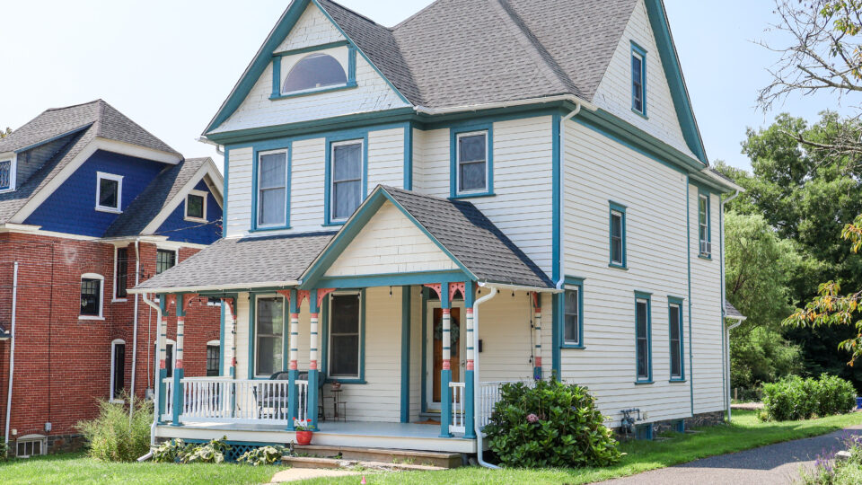 Two-story cream house with teal trim and wraparound porch beside a brick home on a sunny street.