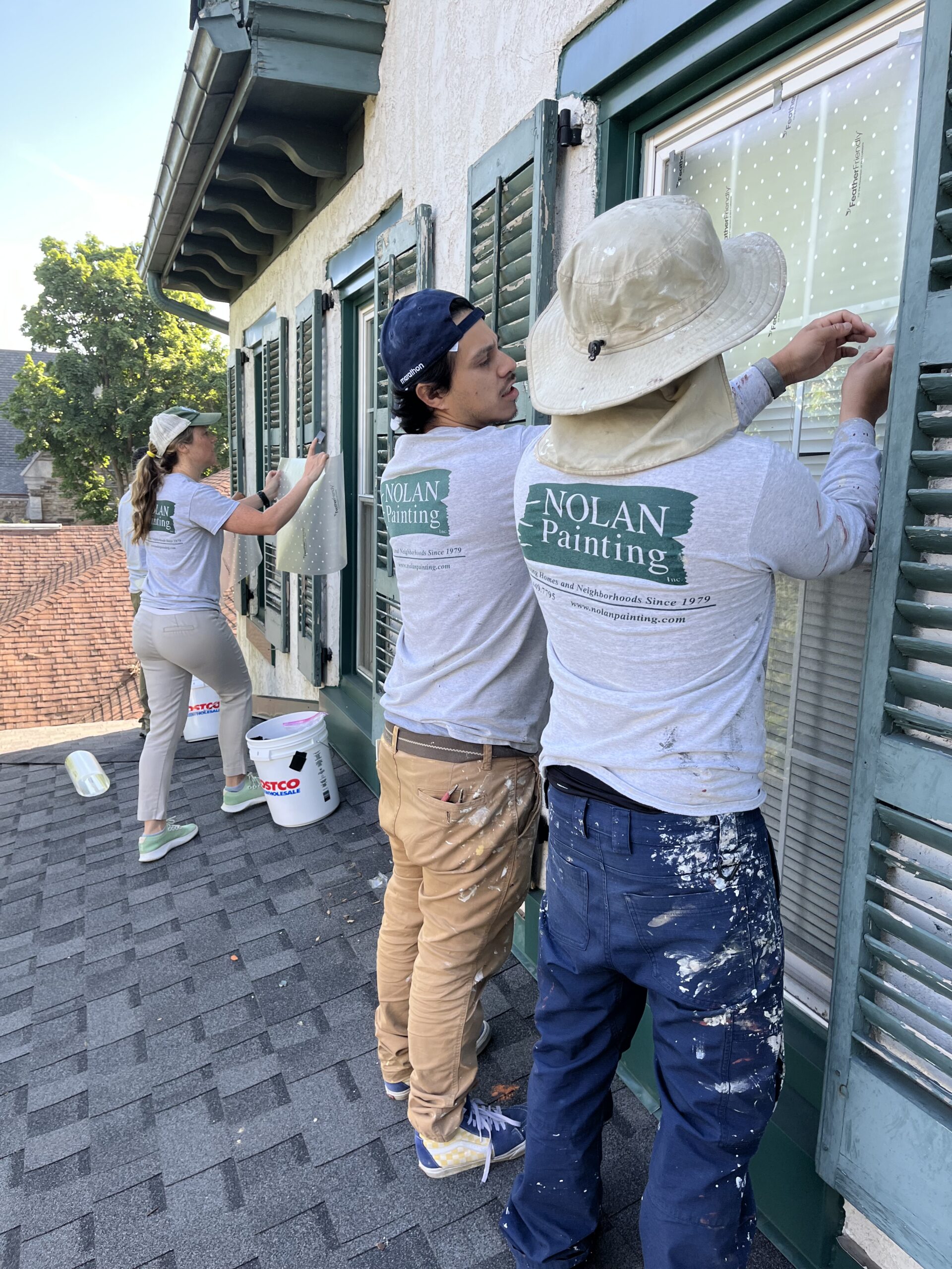 Nolan Painting Company- Havertown, PA Three painters on a roof working on exterior windows, taping protective plastic sheets over a green-framed wall; all wear Nolan Painting shirts and paint-splattered clothing.