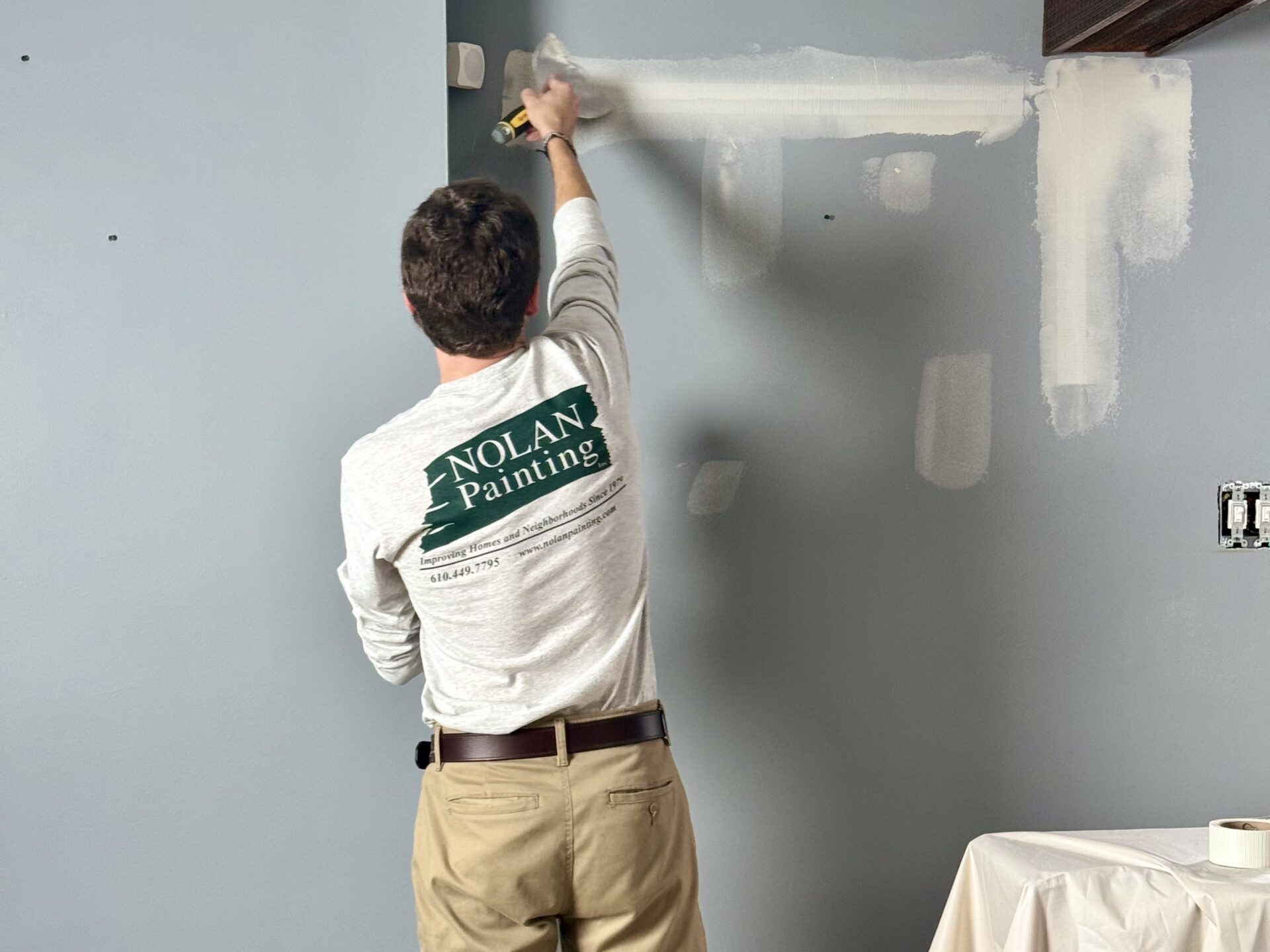 Worker painting a blue-gray wall using a paint roller, wearing a Nolan Painting shirt and khaki pants in a home interior.