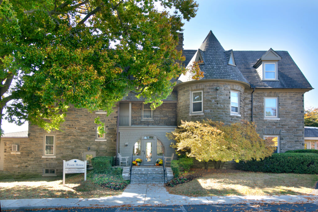 Front view of Tyson House: a stone mansion with a rounded turret, slate roof, and leafy trees in the foreground; a Tyson House sign sits near the entrance.