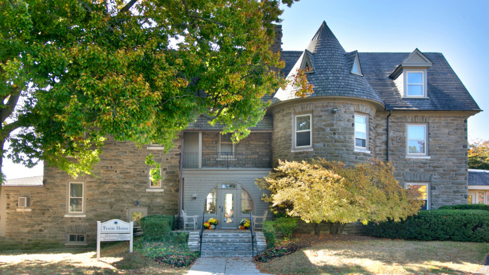 Front view of Tyson House: a stone mansion with a rounded turret, slate roof, and leafy trees in the foreground; a Tyson House sign sits near the entrance.