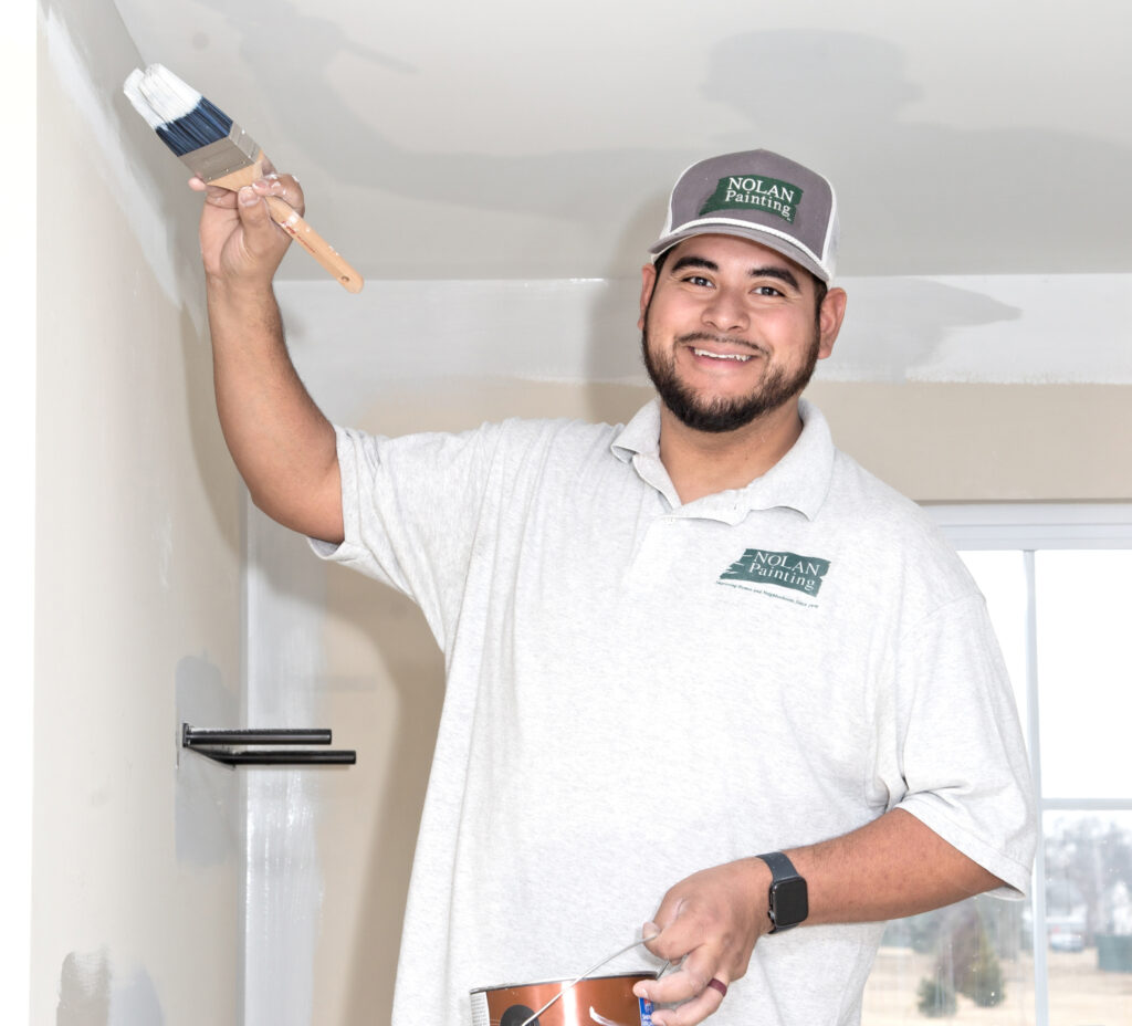 Smiling painter in a gray Nolan Painting cap brushes white paint onto an interior wall with a wide brush.