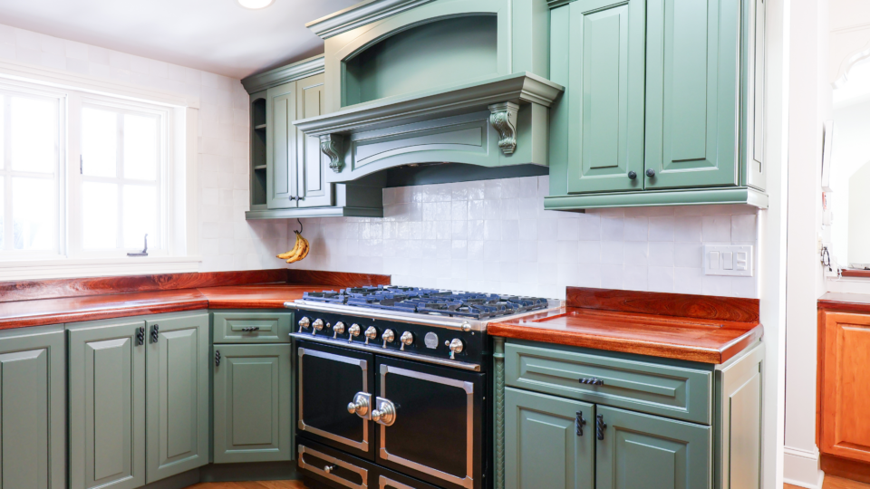 Green cabinetry surrounds a stainless steel pro-style gas range with double ovens and wooden countertops, in a bright kitchen. Banana hangs on the wall nearby.